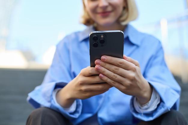 Primer plano retrato de una mujer joven sosteniendo un teléfono inteligente en ambas manos sentada en la calle con el móvil